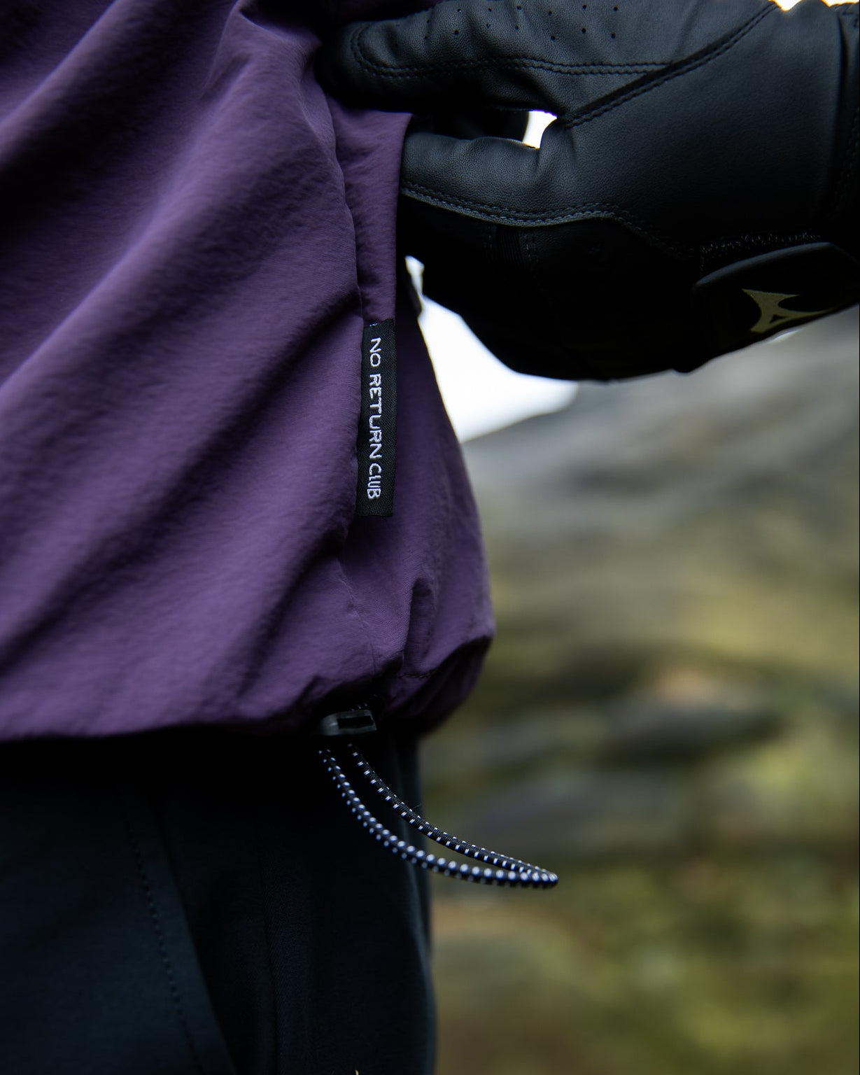 Person wearing a purple jacket with a brand label, holding the hood, against a blurred natural background.